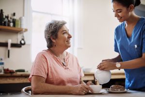Caregiver, smile and woman with tea in nursing home kitchen for breakfast, relax or healthy nutrition of support. Nurse, patient and food for senior care with helping, trust or serving at retirement.
