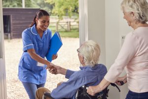 Senior Couple With Man In Wheelchair Greeting Female Nurse Or Care Worker Making Home Visit At Door